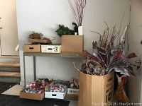 View of metal shelves and floor with multiple boxes and a large cardboard box containing faux flowers, pine branches, and crafting supplies.