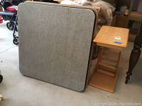 Two gray padded folding card tables stacked next to a wooden side table with magazine rack shown on concrete floor in garage setting.