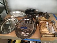 Silverplate serving ware including platters, bowls, pitcher and boxed presidential spoons on metal shelving.
