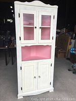Front view of tall white corner display cabinet with upper glass doors, a middle open shelf, and lower solid doors.