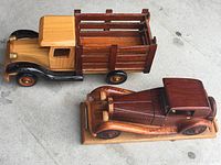 Two hand-carved wooden vehicle models displayed together on a concrete floor, showing the pickup truck with slatted wooden sides and a darker stained 1935 Mercedes Benz model on a wooden base