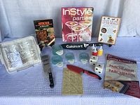 Photo of kitchenware and cookbooks showing knives, salt and pepper dice shakers, miniature condiment containers, parchment paper, microwave bacon cooking tray in original box, and cookbooks.