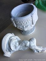 Photo of decorative outdoor cement flower pot and plaster cherub wall mount side by side on a table.