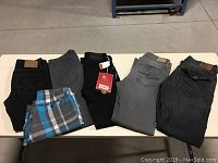 Six pairs of pants and shorts laid out on table showing black, grey, dark grey jeans and one blue plaid shorts.