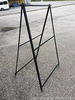 Black metal sign frame standing upright on pavement, showing foldable A-frame structure and metal crossbars.
