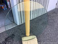 Full view of large circular glass tabletop leaning against a yellow pole outdoors, showing overall condition and size.
