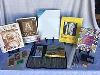 Lot of artist supplies including paint brushes, palettes, various paper pads, oil pastels, pencils, canvases, and craft items laid out on a grey tablecloth.