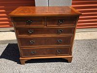 Front view of vintage wooden chest showing 5 drawers with metal handles and bracket feet, placed on asphalt ground.