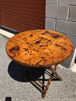 Round bamboo folding table positioned near a concrete block wall and red metal door, showing the top surface of the table with distinctive dark spotting and wood grain pattern.