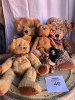 Photo showing all seven bears arranged on a decorative table with a MaxSold lot number card present, including the Ty Beanie Baby bear with visible tag.