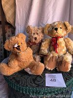 Three tan-colored collectible teddy bears are sitting on a black fringed cloth on a round surface. One wears a red ribbon, and one holds a toy bee.