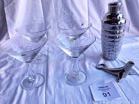 Four heavy crystal martini glasses and silver cocktail shaker with bottle opener on white cloth background.