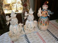 Three Jim Shore snowmen figurines displayed on a table, showing detailed hand-painted designs and small animals like owls and dogs.