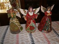 Front view of three angel figurines on a patterned tablecloth showing detailed hand-painted designs and white wings.