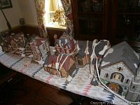 Side view of eight miniature Dickens village houses lined up on a patterned cloth, showing detailed roofs with artificial snow, chimneys, and brick textures. Electrical cords attached to some.