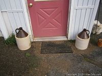 Wide shot showing both antique stoneware jugs with natural glazed bodies and dark brown Albany slip tops positioned outside near a door.