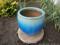 Photo of the large blue/green round ceramic pot sitting on a concrete base outdoors among plants.