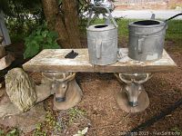 Bench composed of wooden seat with two deer head supports and cement lion beside it, with two metal watering cans resting on the bench.