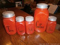 Front view of all five red kitchen storage jars with labels Tea, Sugar, Salt, Coffee, and Flour arranged on wooden table.