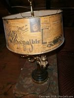 Side view of the vintage lamp showing the brass fixture, dark metal base, and the unique lampshade covered in old newspaper clippings with various texts and pictures.