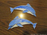 Two blue stained glass dolphins laid on a wooden surface, showing full shapes and color variations