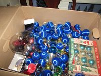 Open cardboard box filled with large blue velvet balls, some silver balls, vintage boxed ornaments, and a clear container with small colored balls.