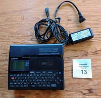 Black Casio CW-K85 disc title printer with QWERTY keyboard, LCD display and function buttons with power cord and extra ink cartridge.