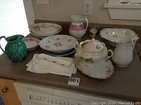 Collection of decorative plates, pitchers, bowls, and covered sugar bowl arranged on countertop, showcasing floral patterns and vintage styles.