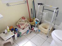 Wide view of bathroom assistance items, cleaning supplies, and household items arranged on tiled floor near wall.