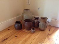 Six pottery jars and a pitcher placed on wooden floor corner, showing variety of sizes and rustic glaze.