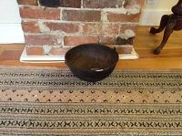 Cast iron bowl with spout and handle displayed on a patterned rug in front of a brick fireplace.