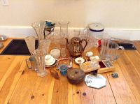 Wide view of all items in the lot arranged on wooden floor including glass vases, colored glass shoe and hat, pitchers, tea cup, ash tray, flour tin, and various decor pieces