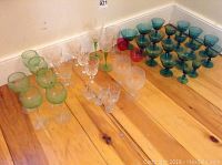 Wide shot of the mixed stemware collection showing green-tinted, clear crystal, cranberry, and blue tinted glasses arranged on wooden floor.