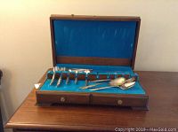 Wooden canteen box opened showing blue velvet inner lining with assorted mismatched silver-plate utensils including forks, spoons, and knives.