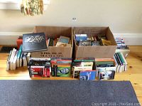 Two cardboard boxes filled with books on a wooden floor with some books visible by their spines and covers