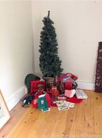 Photo showing the artificial Christmas tree in planter with various Christmas holiday items arranged around it including gift bags and boxes, towels, teddy bear, and tree stand.