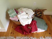 Wicker basket filled with folded white linens, pastel towels, and a red lambswool blanket placed beside a basket of wooden hangers on a wood floor.