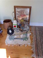 Full shot of lot items: floor mat with animal designs and fringed edges, vintage stuffed toys (bunny with stained jacket, dog with yellow hat, bear in blue dress), plastic black police hat with silver badge, white lamp with red apple design base, and framed 'Anatomy of a Teddy Bear' print leaning on wall.
