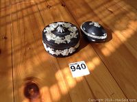 Two Wedgwood black jasperware dishes with white relief decorations sitting on a wooden table, one egg-shaped and one round with lid.