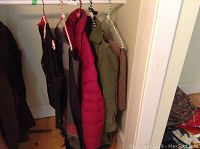 Clothing items hanging on a rack: dark suede vest, grey vest, red goose down jacket, olive green wool jacket and brown corduroy dress or skirt.