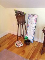 Full view of tall wicker plant stand with basket top and colorful liner, brown plant pot on floor, gardening books stacked on floor, and tall floral garden plaque poster leaning against wall