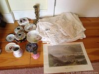 Overview of lot items on floor with tablecloths, dishes, candle holder, and prints.