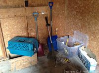 Photo showing blue plastic pet carrier on wooden box, two shovels, snow shovel, plastic bin with assorted items, and a purple shovel, all inside a storage shed.