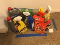 Photo showing assortment of household cleaning supplies including spray bottles, jugs, glove bags, and aerosol cans on a carpeted floor.