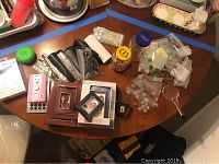 Full view of assorted items on oval wooden table including Bluetooth earbuds in box, pink device, picture frames, currency, glasses, and small containers.