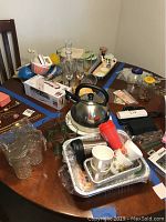 Overall view of all kitchen items on wooden table showing glasses, kettle, utensils, trays, mugs, and more.