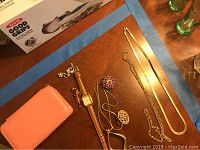 Jewelry lot including chains, brooches, watch, and pink fabric jewelry box displayed on a wooden surface with blue painter's tape borders visible.