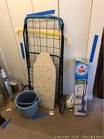Wide shot showing full height black metal handcart, ironing board with floral cover leaning on it, boxed Bissell vacuum, blue bucket with smaller bucket inside it on the floor in front, and small blue wastebasket.