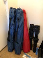 Four large folding camping chairs in blue and red carry bags and two folded smaller black folding chairs leaning against a wall.