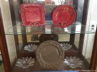 Set of five items displayed on glass shelf: two red decorative plates upright on metal stands, one large brown plate lying flat, and two flower-shaped pressed glass dishes.